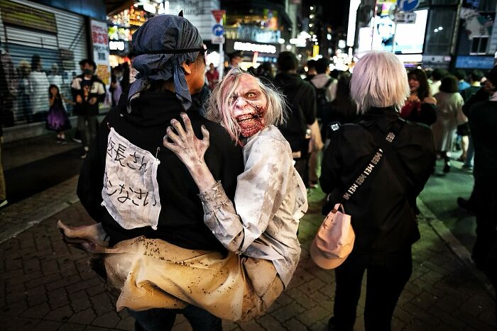 Street photo capturing humanity and absurdity with a person carrying a zombie-like figure in a busy nighttime urban setting.