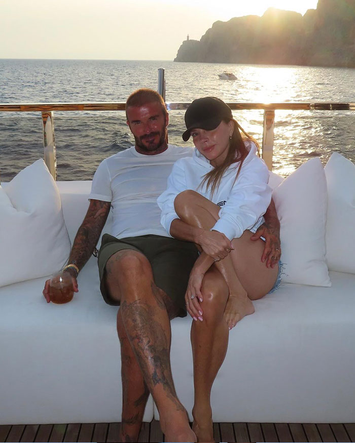 David Beckham and a woman sitting on a white couch on a boat with ocean and cliffs in the background at sunset.