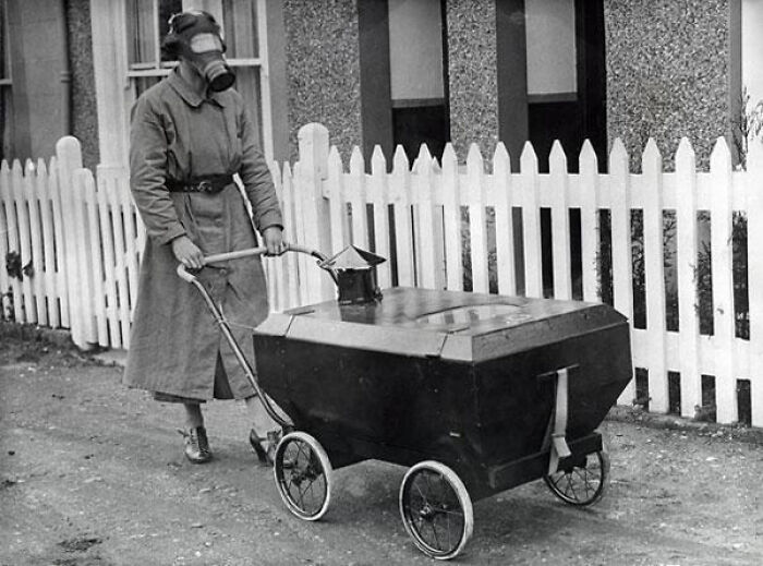 Person wearing a gas mask pushes a strange, boxy stroller on a sidewalk in an old black and white creepy photo.