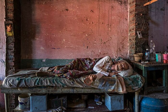 Elderly man resting on a worn bed in a humble room, capturing candid street photo of humanity and tender moments.