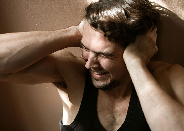 Man in a black tank top grimacing with hands on his head, expressing stress in a dimly lit room, revenge acts theme.