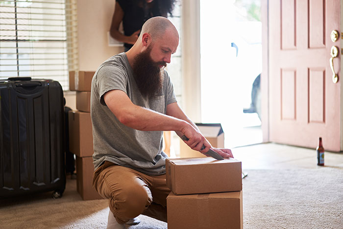 Bearded dad opening packed boxes at home, reflecting on life changes after difficult accusations from his kids.
