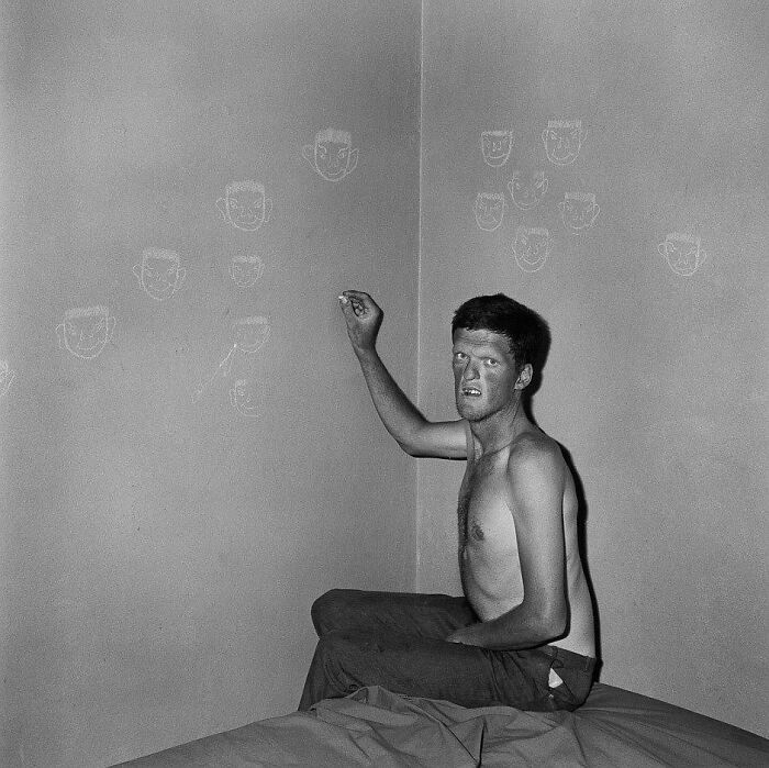 Black and white photo of a shirtless man drawing odd and creepy faces on a wall in a dimly lit room.