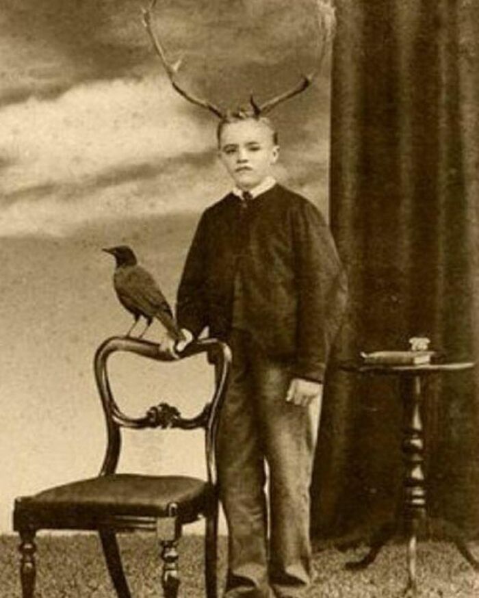 Vintage black and white photo of a boy with antlers standing beside a chair with a bird, odd and creepy imagery.