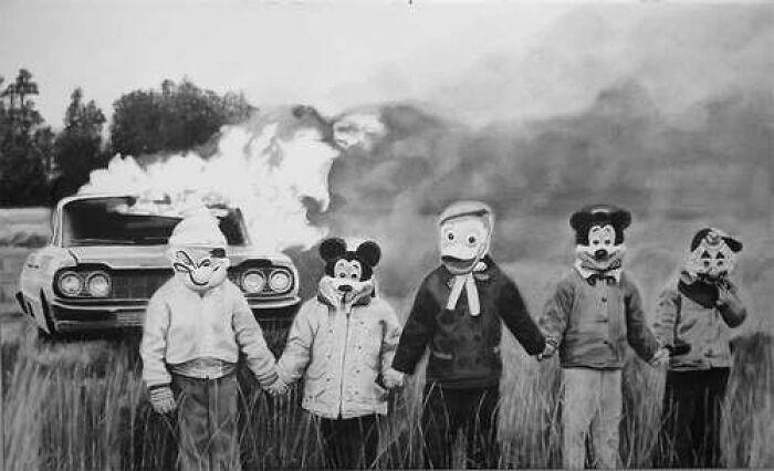 Five children wearing eerie vintage cartoon masks hold hands in a field with a car engulfed in flames behind them in a creepy old black and white pic.