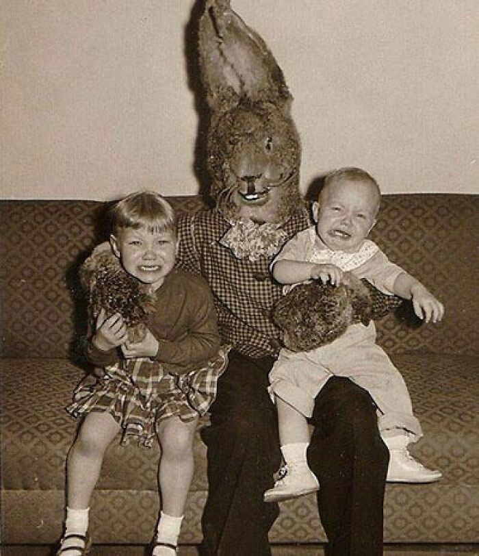 Vintage black and white photo of two children with a person in a creepy rabbit costume on a couch.