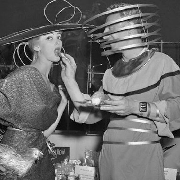Black and white photo of two people in odd and creepy vintage costumes, one feeding the other with unusual headgear.
