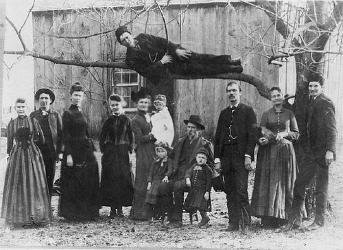 Old black and white photo of a vintage family portrait with a man lying oddly on a tree branch, adding a creepy vibe