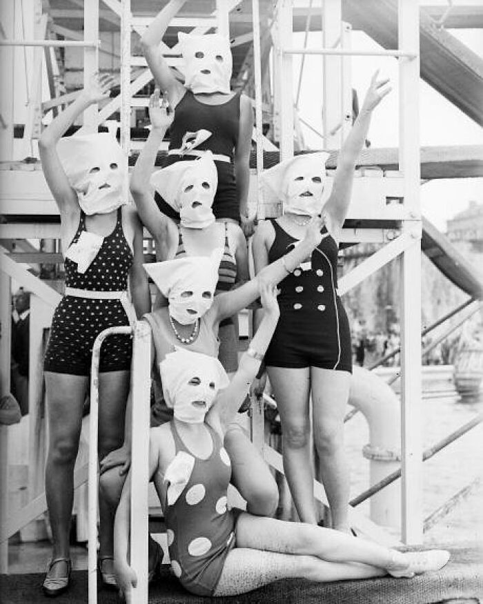 Group of women in vintage swimsuits wearing odd and creepy masks posing on wooden structure in an old black and white pic.