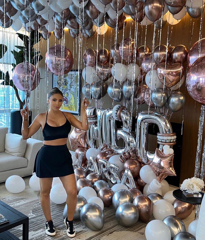 Larsa Pippen posing in black outfit surrounded by silver and rose gold balloons for plastic surgery transformation celebration