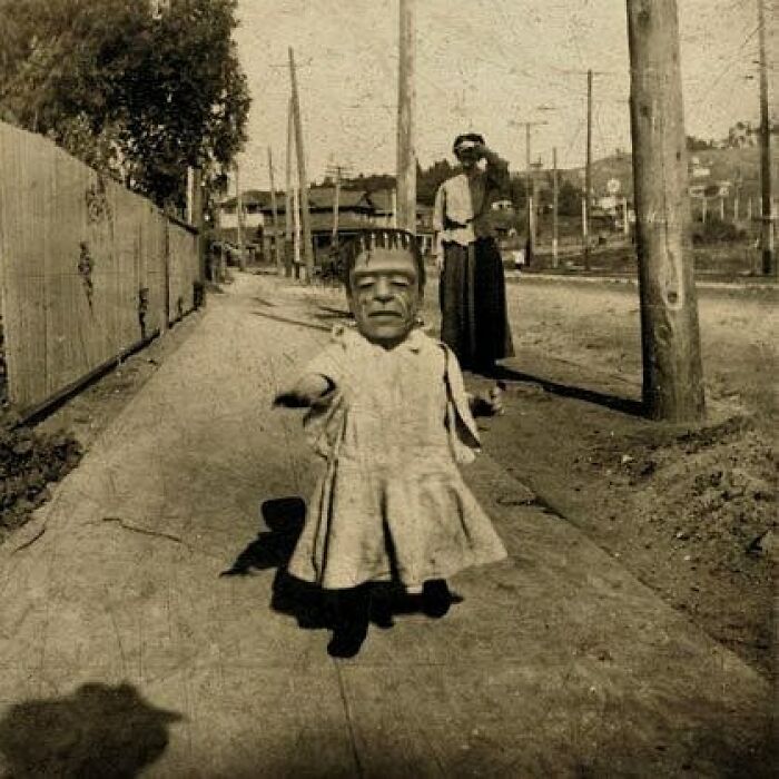 Sepia-toned old black and white photo featuring a child with a Frankenstein mask on a sidewalk, eerie and odd vibe.