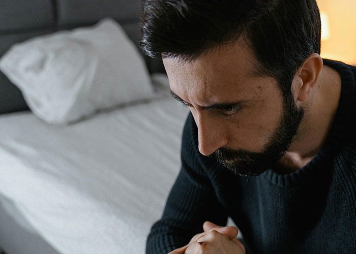 Man with beard sitting on bed, appearing deep in thought, reflecting on life changes after learning about biological parentage.
