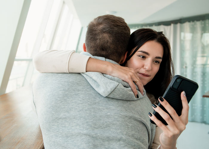 Woman hugging partner while checking phone, tense expression suggesting dating deal-breakers and trust concerns.