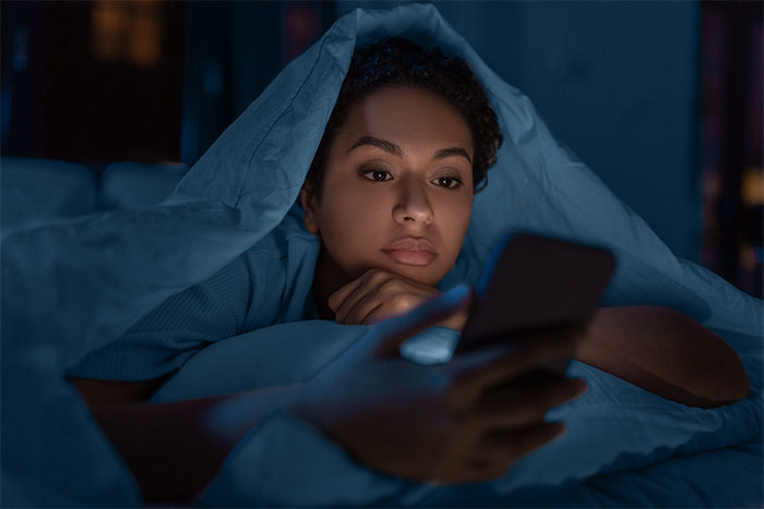 Woman lying in bed under covers, focused on her phone screen late at night, reflecting on coworker situation.