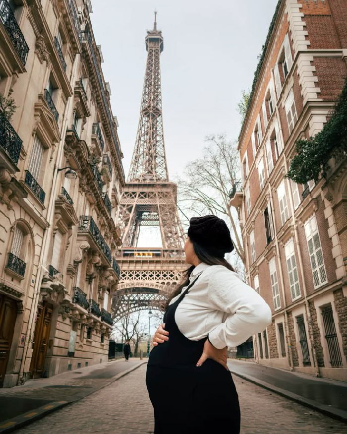 Pregnant woman posing by the Eiffel Tower in Paris, holding her belly, maternity photoshoot ideas.