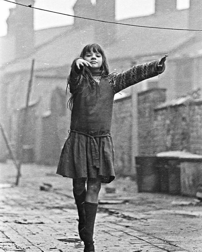 Black and white photograph of a young girl walking in a worn urban street, capturing a moment of humanity’s sad and beautiful history.