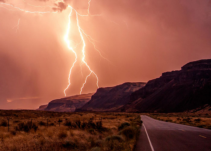 Lightning striking over a deserted road with mountains in the background, illustrating wacky stories and unusual events.