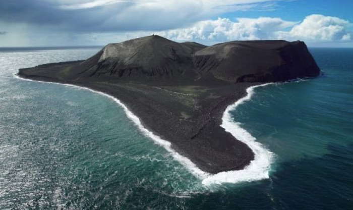 Aerial view of a forbidden place on earth, a remote volcanic island surrounded by ocean waves and cloudy skies.