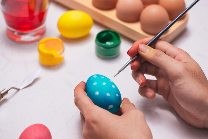 Hands painting a blue Easter egg with white dots, surrounded by colorful paints and unpainted eggs on a white surface.
