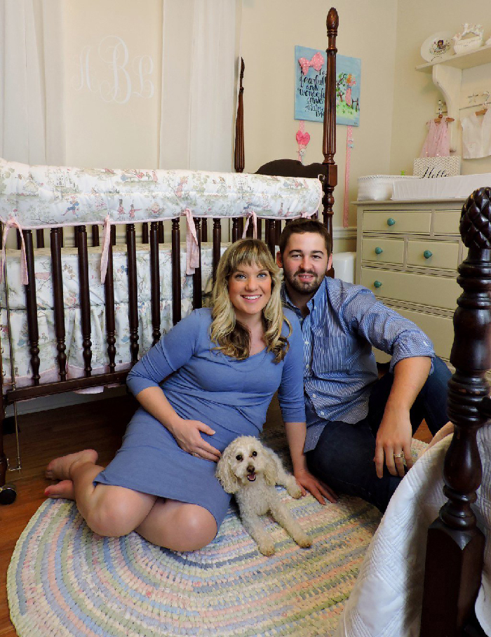 Couple in pastel nursery with small dog; pregnant woman holding belly in maternity photoshoot