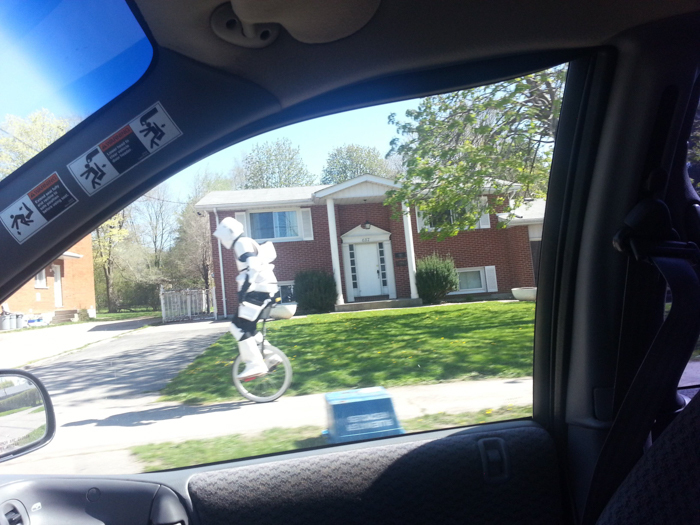 Person dressed as a stormtrooper riding a unicycle on a suburban sidewalk, captured through a car window.