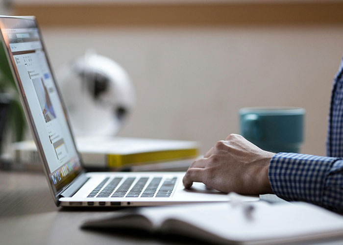 Person in a checkered shirt typing on a laptop, researching perfect revenge acts that never got caught at a desk.
