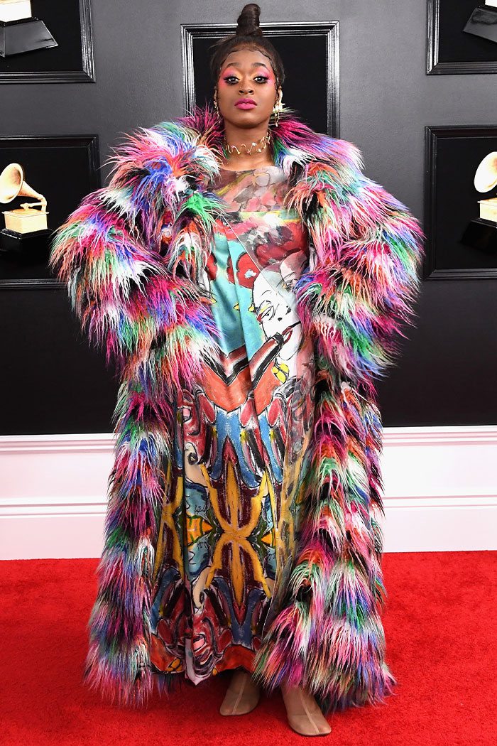 Woman in a colorful, wild fur coat and patterned dress posing on the red carpet at the Grammys with bold makeup and jewelry.