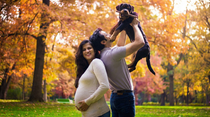 Couple in autumn park during creative maternity photoshoot, pregnant woman smiling while partner lifts dog, colorful leaves.