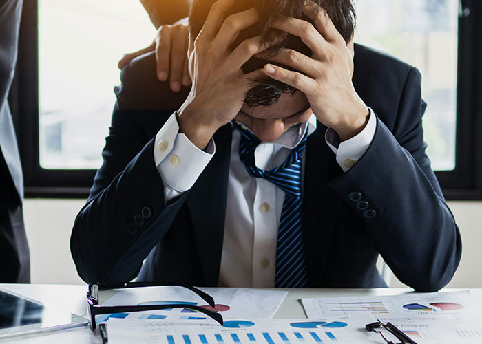 Stressed businessman holding his head with hands at office desk showing frustration after perfect revenge acts.
