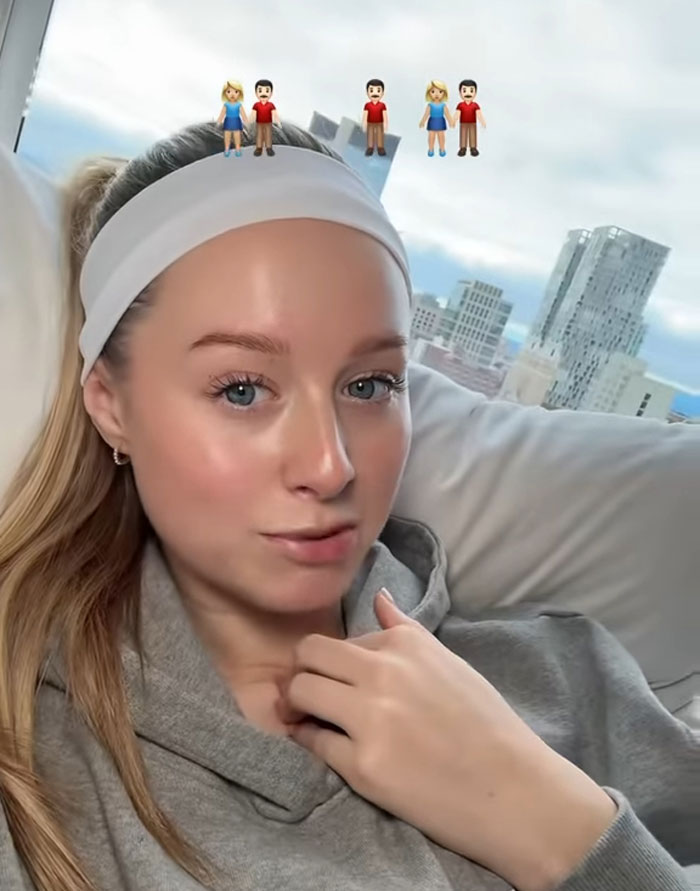 Young woman sharing why she has 7 parents, sitting indoors with emojis of multiple parents above her head.