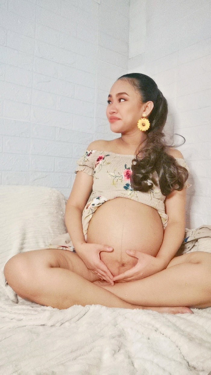 Pregnant woman seated cross-legged, hands forming a heart on belly in floral top for a maternity photoshoot