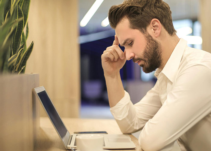 Young man with beard and white shirt looking stressed and thoughtful while working on a laptop indoors, reflecting neurodivergent habits.