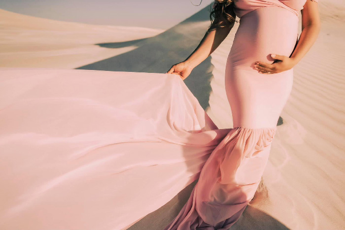Pregnant woman in pink flowing gown on sand dunes, holding belly, maternity photoshoot ideas with long train and soft light