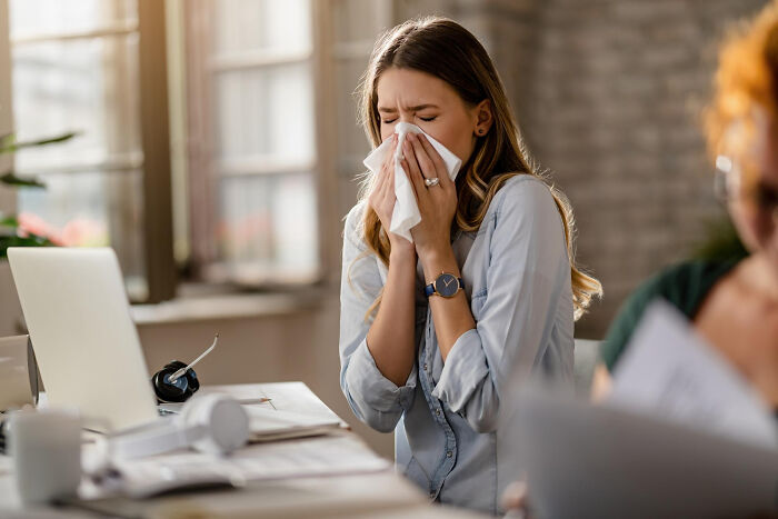 Young woman sneezing into tissue at office desk, experiencing moments that hit people harder than expected.