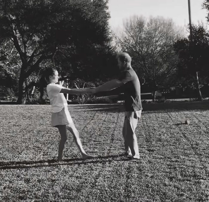 Meghan Markle and Prince Harry holding hands and spinning in a sunny outdoor park with trees in the background.