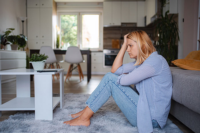 Upset woman sitting on living room floor, holding her head, portraying embarrasses wife moment and domestic tension