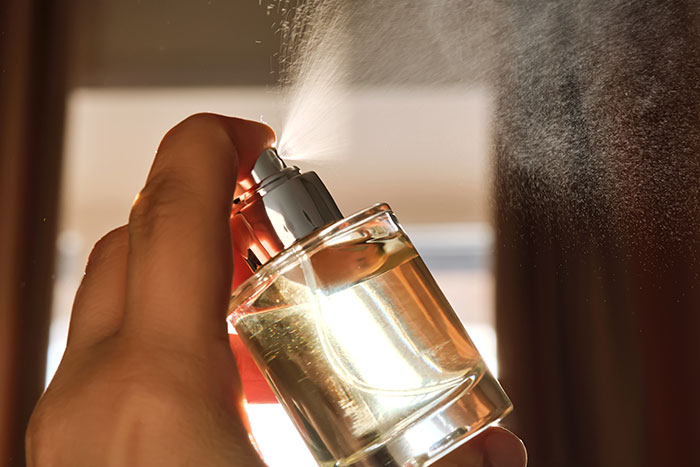 Hand spraying perfume from a clear glass bottle with mist visible in warm indoor lighting, MIL runs to the priest concept.