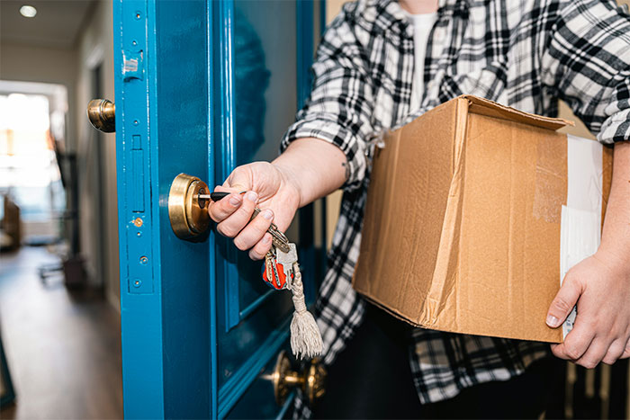 Person holding a cardboard box unlocking a blue door, symbolizing moving in next door to a former friend.