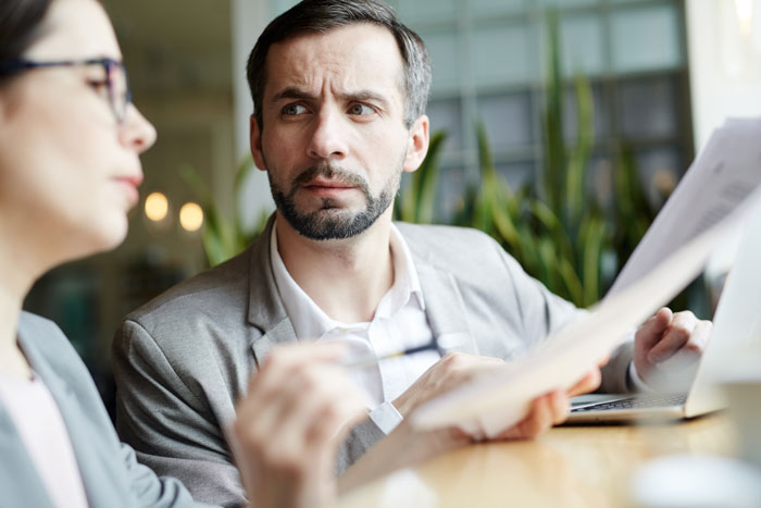 Man in a gray suit looking puzzled while a female coworker talks during a tense office interaction.