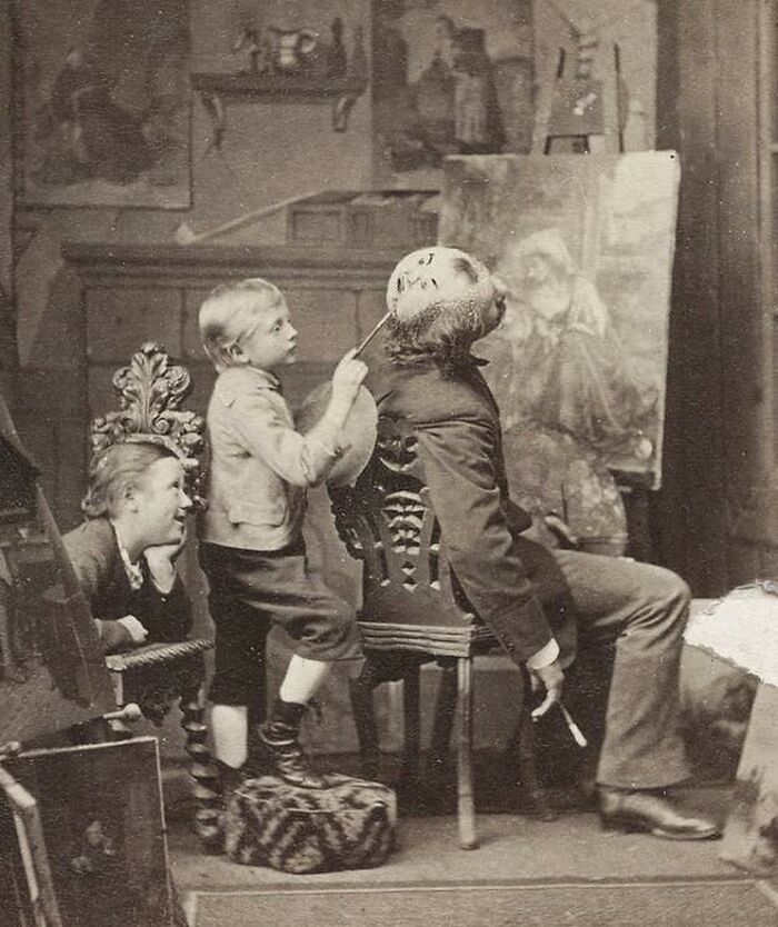 Old black and white creepy photo of a child painting an eerie face on a seated man's head in a vintage room.