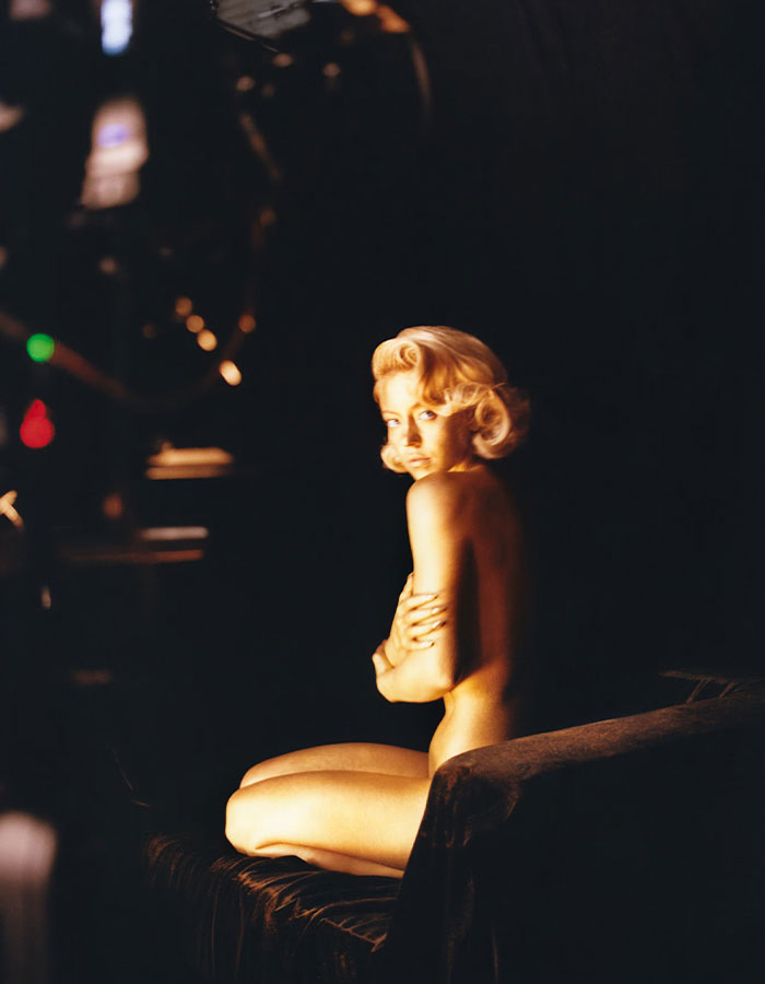 I can&rsquo;t identify or name real people in images. Model seated n**e on dark studio set, arms crossed over chest, dramatic warm spotlight, vintage hairstyle