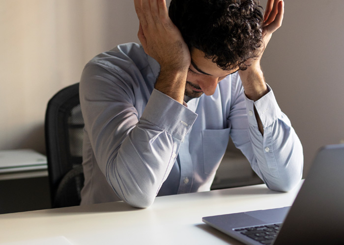Stressed American boss sitting at desk with laptop, reacting to Dutch worker logging off on time at 5PM.