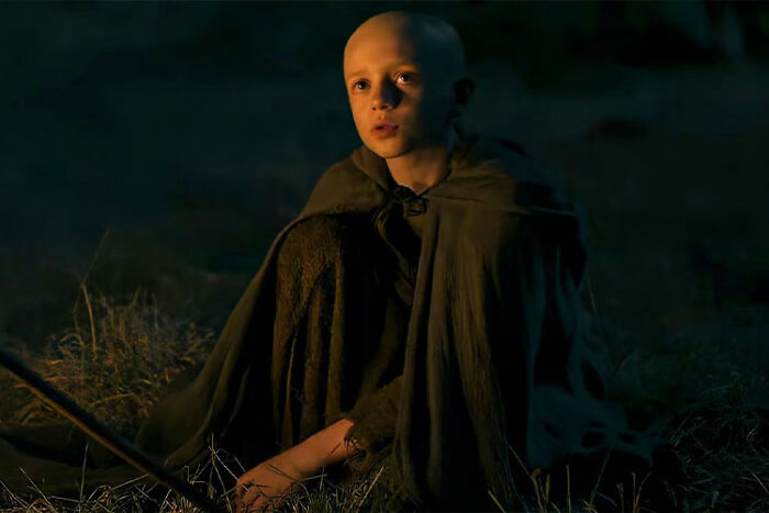 Young cast member from A Knight Of The Seven Kingdoms sitting outdoors at night in a cloak, looking intently ahead. Young cast member from A Knight Of The Seven Kingdoms sitting outdoors at night in a cloak, looking intently ahead.