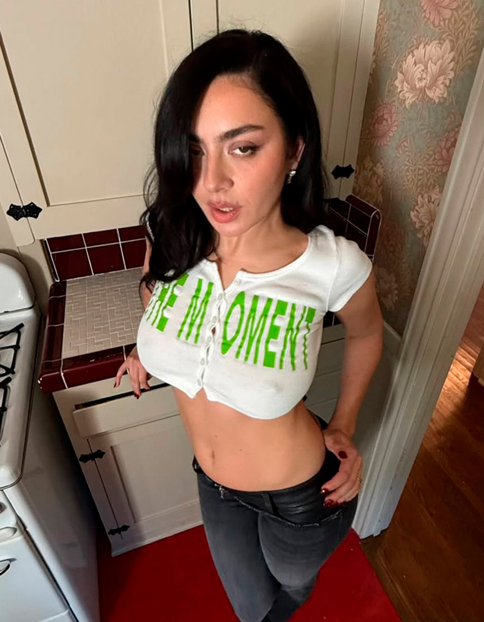 Charli XCX posing braless in a cropped button-up top and black pants in a vintage-style kitchen setting.