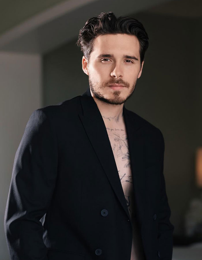 Young man with tattoos wearing an open black blazer, representing footage of Victoria Beckham after Brooklyn's dance claims.