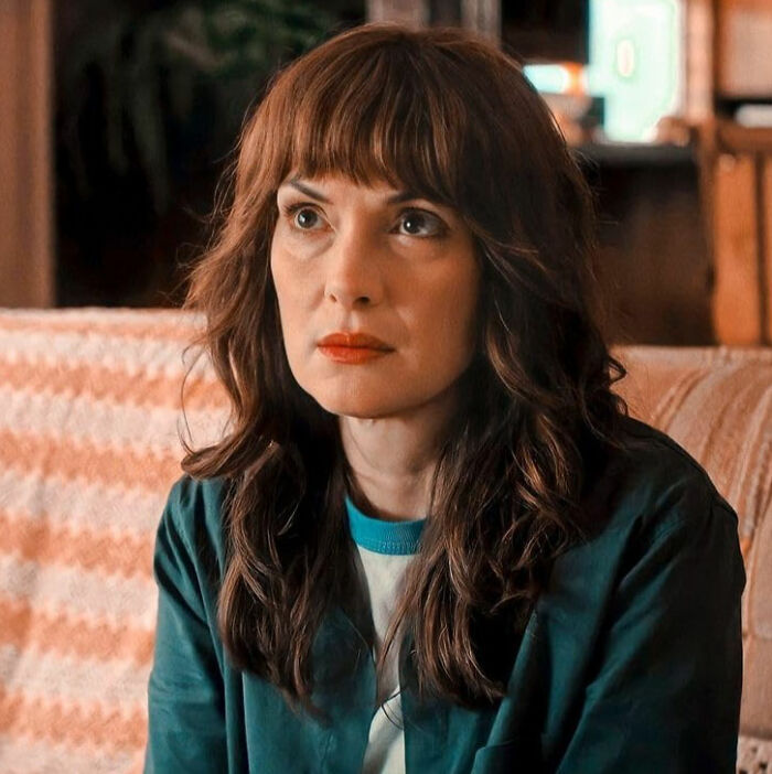 Woman with brown hair and bangs, wearing a teal jacket, portraying a celebrity downfall with a serious expression indoors.