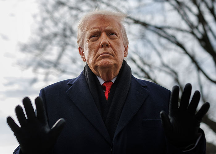 Man in a dark coat and gloves raising his hands outdoors with bare trees in the background during winter.
