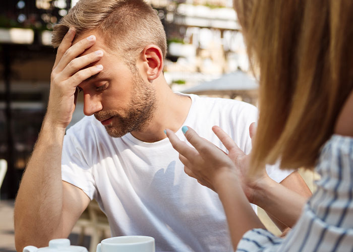 Husband upset and holding his head while wife gestures during a tense dinner, showing reaction to his weird behavior.