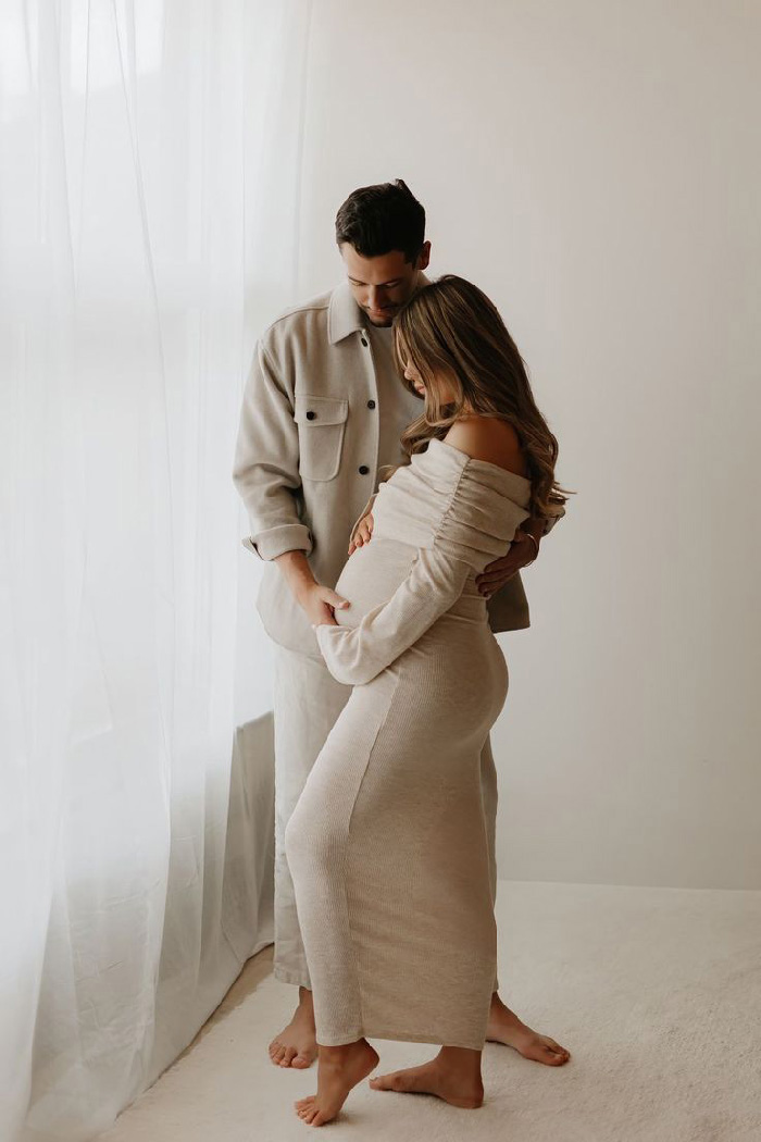 Couple embracing by sheer window, pregnant woman in beige dress — maternity photoshoot ideas with soft natural light
