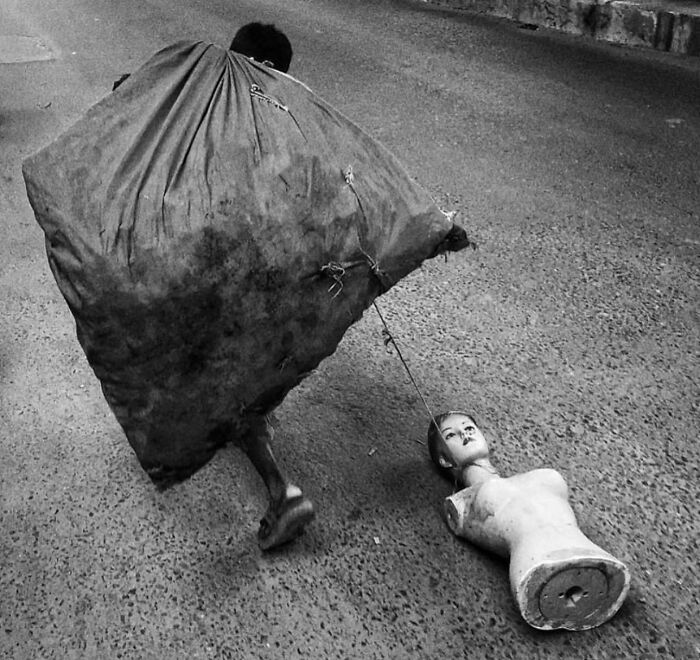 Man carries a large sack while pulling a mannequin head attached by string in a striking street photo capturing unexpected moments.